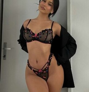 Woman in black sheer lingerie with pink floral embroidery posing in a doorway, robe draped over her shoulders in a light room Woman in black sheer lingerie with pink floral embroidery posing in a doorway, robe draped over her shoulders in a light room
