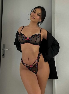 Woman in black sheer lingerie with pink floral embroidery posing in a doorway, robe draped over her shoulders in a light room